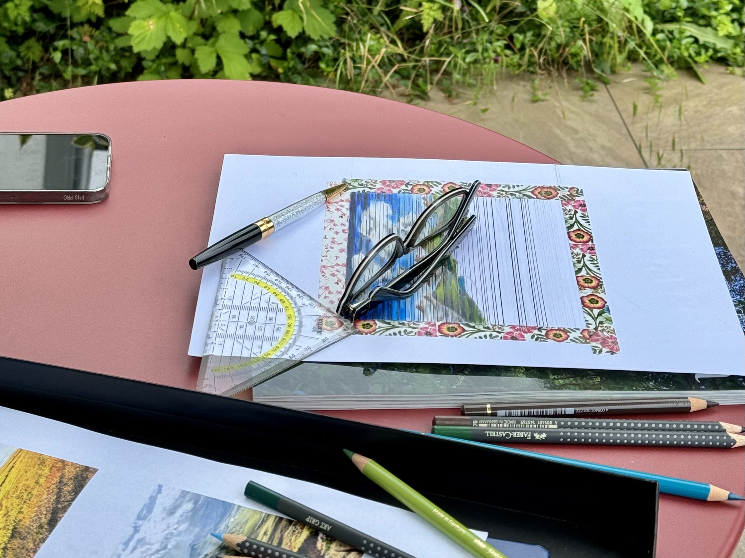 Ein Tischchen mit Zeichnungsutensilien, Lesebrille, Kugelstifte, Blei- und Farbstifte, ein Geodreieck. Eine unvollständige Zeichnung mit einem blumigen Rand. Im Zentrum eine Zeichnung mit horizontalen Streifen. Am Rand des Fotos erkennt man ein Handy und Pflanzen…